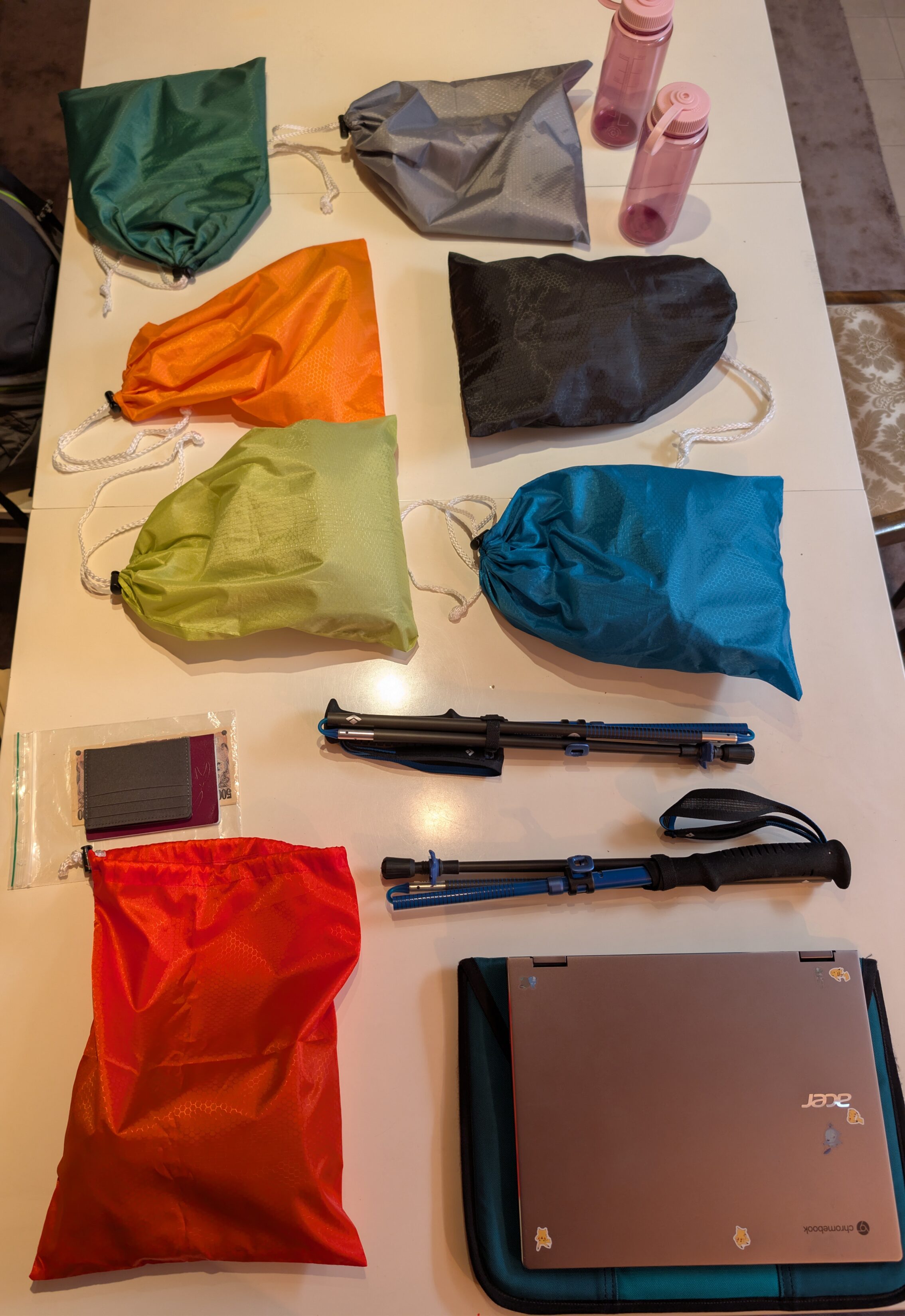 Seven differently coloured packing bags on a table, together with hiking poles, a wallet, a computer, and a couple of water bottles.