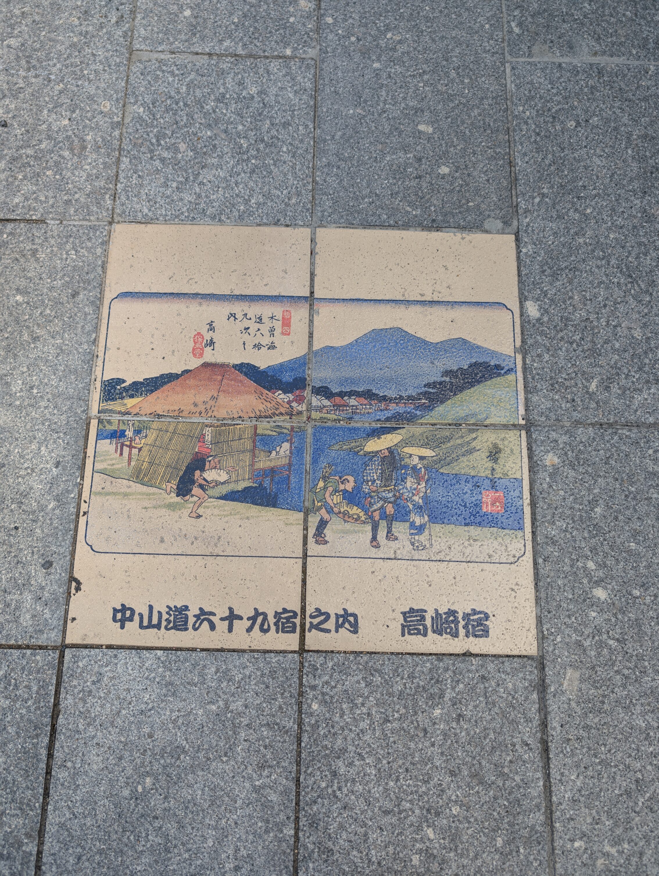 A set of four paevment stones with pictures that together form an 19th century illustration of people walking near a river with mountains in the background.