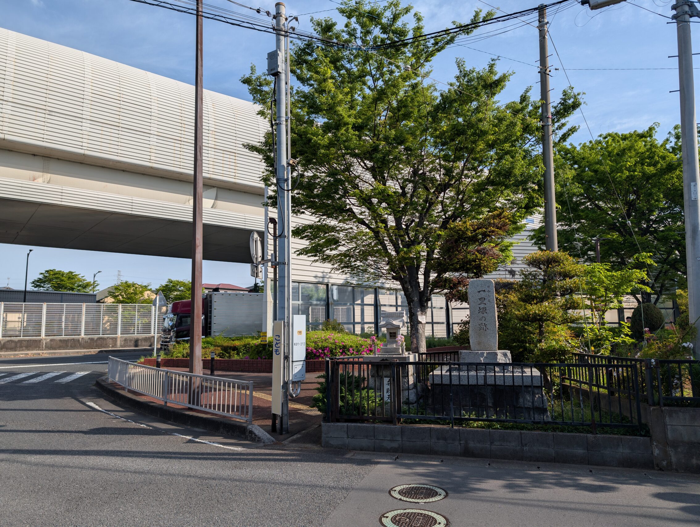 A large tree in front of a huge encapsuled fly-over motorway. In the shade of the tree is a white standing stone.