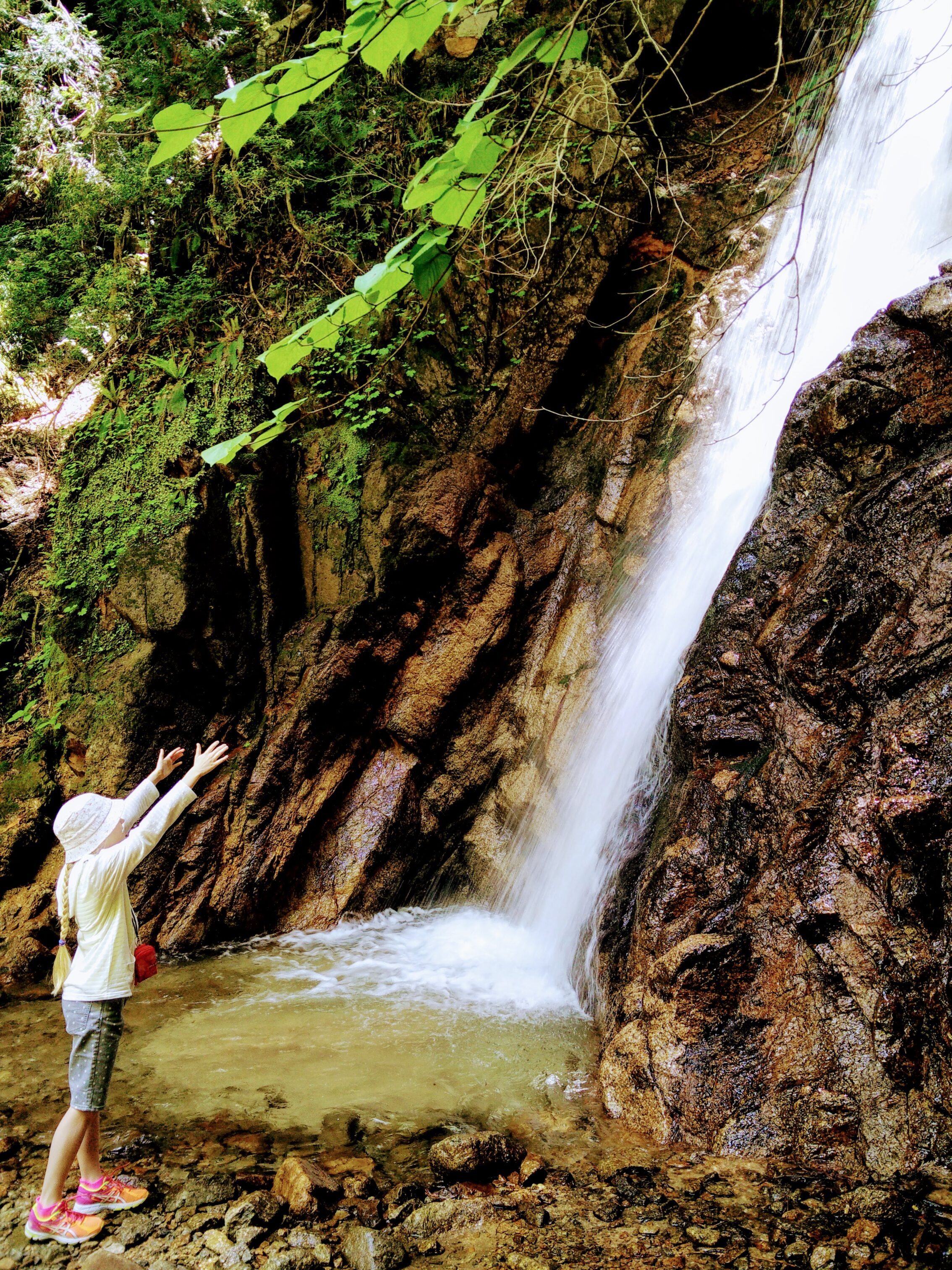 A young girl in a white hat is stretching her arms towards a waterfall.