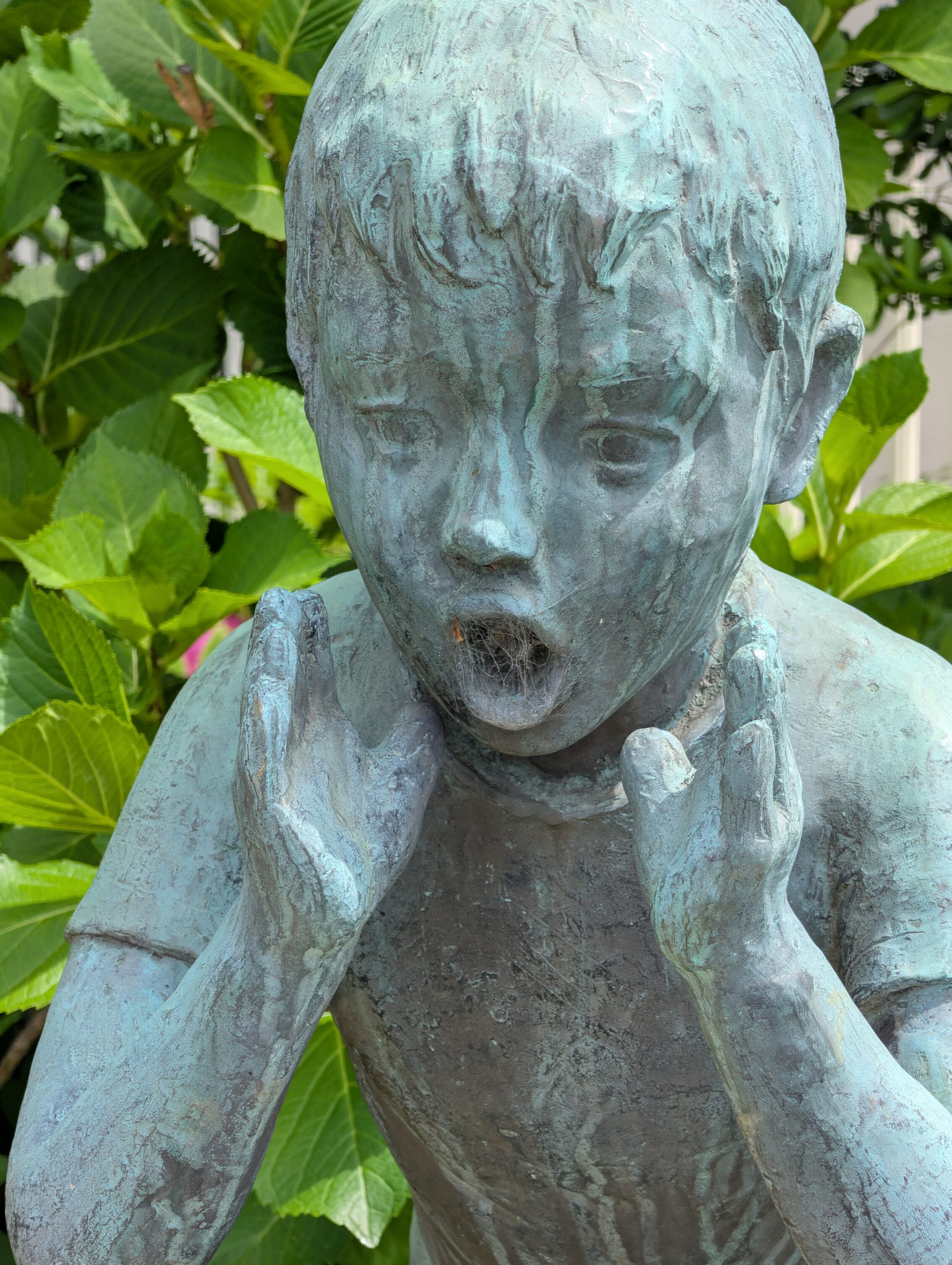 A closeup of a copper statue of a shouting boy, with cobwebs covering his mouth.