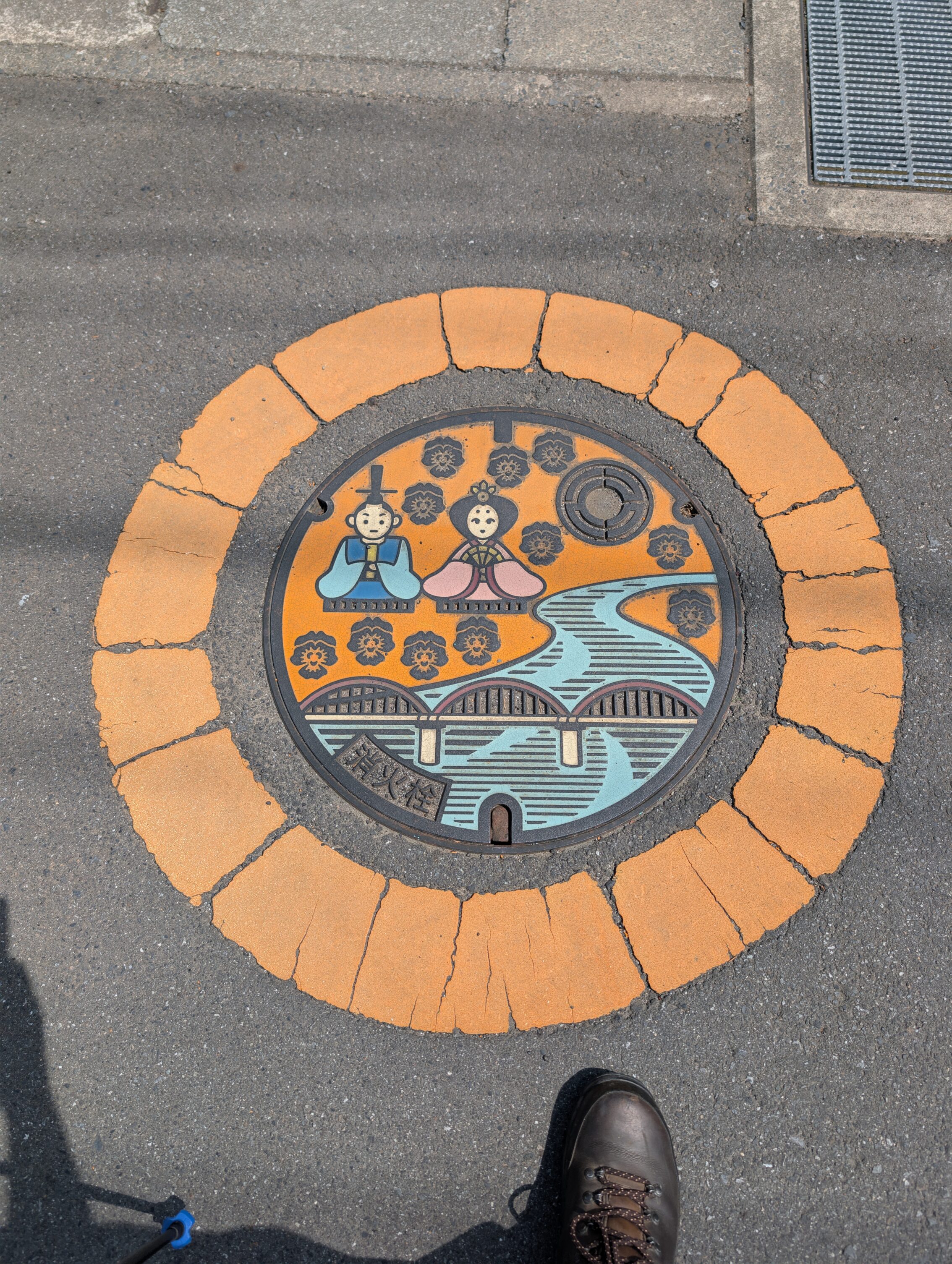 Manhole cover featuring two people dressed in traditional clothing with a river, and a modern bridge.
