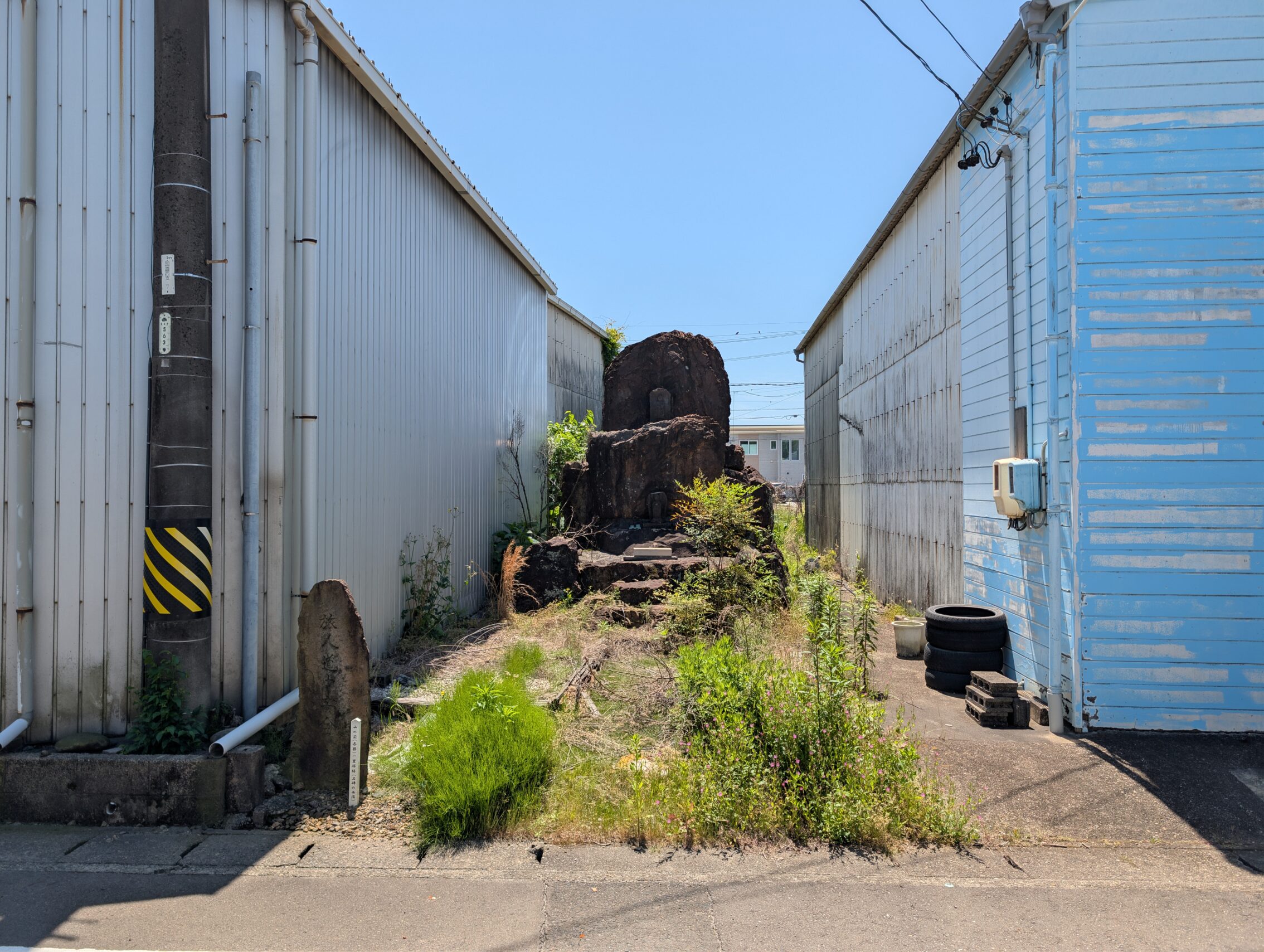 Two warehouse buildings with a tiny alley between them. In the alley there is a lot of weeds growing, as well the remains of a stone temple.